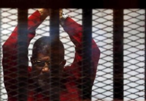 Deposed President Mohamed Mursi greets his lawyers and people from behind bars at a court wearing the red uniform of a prisoner sentenced to death, during his court appearance with Muslim Brotherhood members on the outskirts of Cairo,